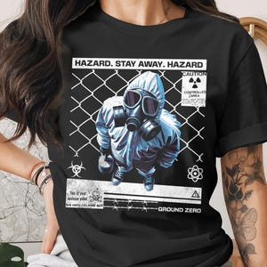 May include: Black t-shirt featuring a person in a hazmat suit and gas mask, standing in front of a chain-link fence. The shirt has the text "HAZARD. STAY AWAY. HAZARD" and other warning symbols and text. The overall theme is a warning.