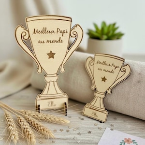 Peut inclure: Deux décorations en forme de trophée en bois. Le plus grand trophée porte l'inscription "Meilleur Papi au monde" et une étoile. Le plus petit trophée porte le même texte et la même étoile. Les deux portent le nom "Ellie". Des épis de blé et des confettis en forme d'étoiles sont éparpillés.