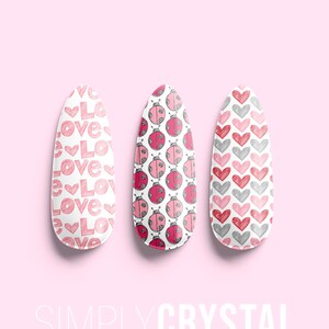 May include: Three oval-shaped nail wraps with different designs. The first wrap has a pink and white pattern of the word "love". The second wrap has a pink and white pattern of ladybugs. The third wrap has a pink and white pattern of hearts. The wraps are on a pink background with the text "SIMPLYCRYSTAL nails" at the bottom.