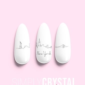 May include: Three white oval-shaped nail tips with black line drawings of the New York City skyline. The middle nail tip has the words "New York" written below the skyline. The text "SIMPLYCRYSTAL nails" is written in red at the bottom of the image.