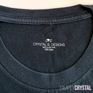 以下が含まれることがあります： 白いタグが付いた黒いTシャツ。タグには「Crystal B. Designs, Made in New York, 100% Cotton」と白い無限大記号が書かれている。タグの下部には「Simply Crystal」と書かれている。
