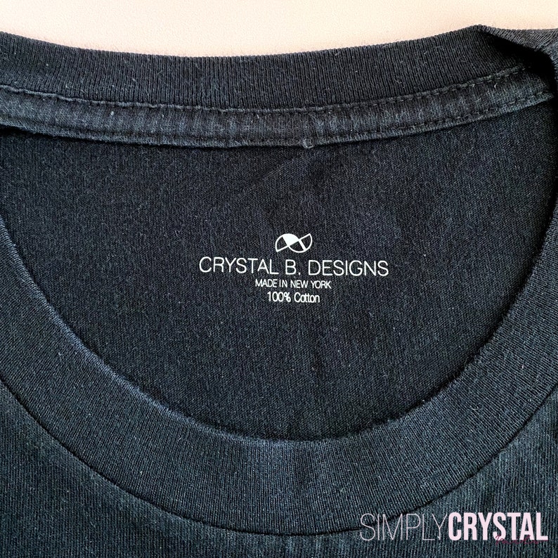 以下が含まれることがあります： 白いタグが付いた黒いTシャツ。タグには「Crystal B. Designs, Made in New York, 100% Cotton」と白い無限大記号が書かれている。タグの下部には「Simply Crystal」と書かれている。