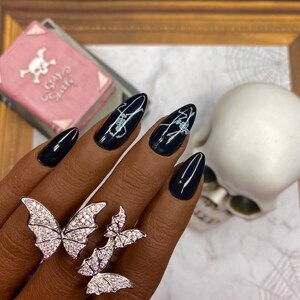 May include: Black stiletto nails with white skeleton designs. A silver ring with three butterfly designs is worn on the ring finger.