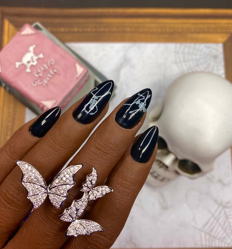 May include: Black stiletto nails with white skeleton designs. A silver ring with three butterfly designs is worn on the ring finger.