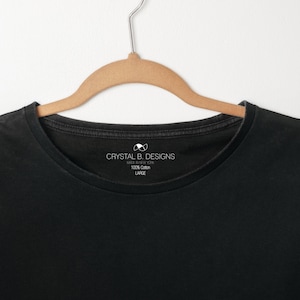 以下が含まれることがあります： 白いタグに「Crystal B. Designs Made in New York 100% Cotton Large」と書いてある黒いTシャツ。Tシャツの下部に「Simply Crystal」の文字が印刷されています。