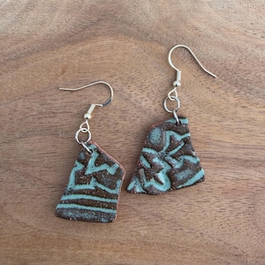 May include: A pair of dangle earrings featuring irregular, flat ceramic shards with a dark brown base and a textured, light blue glaze pattern. Each piece is suspended from a silver-toned metal hook by a small circular jump ring connector.