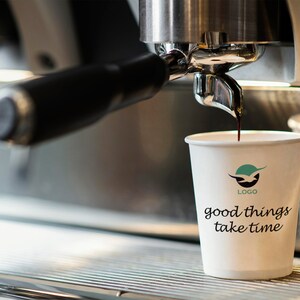 Può includere: Un bicchiere di carta bianco per il caffè con un logo verde e il testo "good things take time". Il caffè viene versato nel bicchiere da una macchina per espresso argentata.