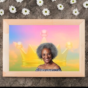 May include: A framed portrait of a smiling woman with gray hair, set against a backdrop of a golden crown and a colorful sky. The frame is made of light-colored wood, and the image is surrounded by white flowers.