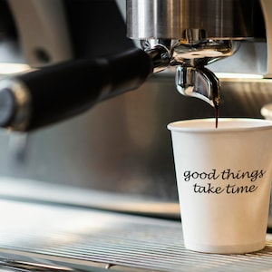 May include: A white paper cup with the text "good things take time" printed on it. Coffee is being poured into the cup from a silver espresso machine.