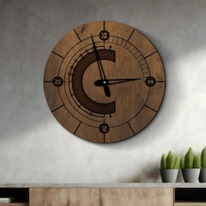 May include: Walnut-coloured wooden wall clock featuring Roman numerals and a large letter "C" at the centre. The clock hands are black. The clock is mounted on a grey wall above a wooden cabinet.
