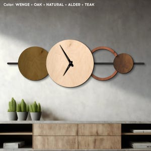 May include: A modern wall clock with a minimalist design. The clock features a large, light-colored wooden circle with black hands, and is accompanied by other wooden shapes in shades of brown and gold. The text at the top says "Color: WENGE + OAK + NATURAL + ALDER + TEAK".