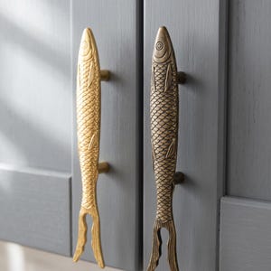 May include: Two vertical cabinet pulls shaped like fish, mounted on gray cabinet doors. One pull is a bright, polished gold tone, while the other is a darker, antique bronze finish. Both feature detailed scale textures and a bifurcated tail design.