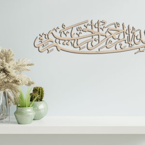 May include: A wooden wall decor with Arabic calligraphy that reads "Bismillah Al Rahman Al Rahim" in a light brown color. The decor is hanging on a light blue wall with a white shelf below it. There are two potted plants on the shelf, one with green leaves and the other with a cactus.