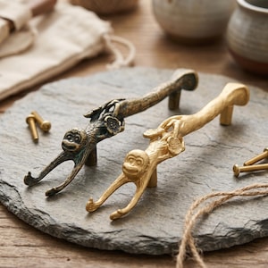 May include: Two metal cabinet pulls shaped like monkeys climbing on branches. One pull has a dark, aged bronze finish, while the other has a bright, polished gold finish. Each handle includes two small metal screws for installation purposes.