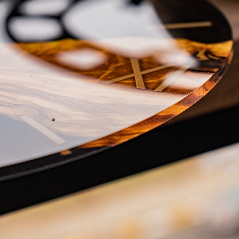 May include: Close-up of a round, wooden clock face with a clear epoxy resin coating. The clock face has a gold-colored design and a black background.