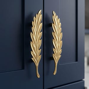May include: Two gold-coloured cabinet handles shaped like elongated leaves with detailed veins and pointed tips. The handles are mounted vertically on a pair of dark navy blue cabinet doors, featuring a smooth, metallic finish and a curved design.