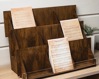 Pode incluir: Um organizador de madeira castanha escura com vários compartimentos, contendo partituras. O organizador tem uma frente de acrílico transparente e foi projetado para exibir e armazenar documentos. A partitura inclui texto como "English Carol Book" e "Composition V5".