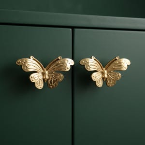 Puede incluir: Dos pomos de armario de metal en tono dorado con forma de mariposa. Cada pomo presenta patrones de alas en relieve detallados con líneas simétricas y pequeños acentos circulares, montados sobre una puerta de armario de color verde oscuro.