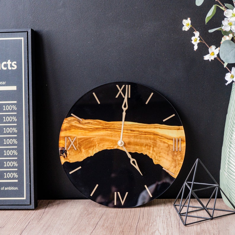 May include: A black and gold clock with Roman numeral markings. The clock face is made of epoxy resin with a wood inlay.