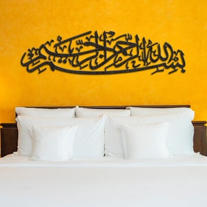 May include: A black wooden wall art piece with Arabic calligraphy on a yellow wall above a white bed with white pillows.