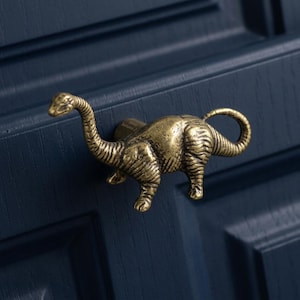 May include: A brass-toned metal cabinet knob shaped like a long-necked dinosaur. The dinosaur figure features a textured, antique-style finish with visible detailing on the neck, body, and tail, mounted horizontally on a dark blue paneled surface.