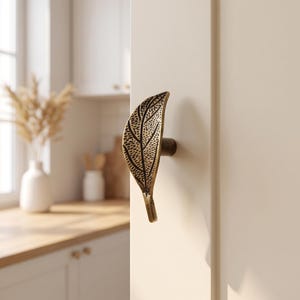 May include: A brass-toned metal cabinet knob shaped like a textured leaf with a central stem and detailed vein patterns. The knob is mounted on a vertical white cabinet door panel, featuring a dark, weathered finish that highlights the leaf texture.