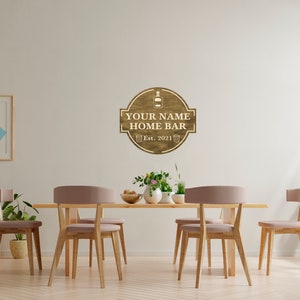 May include: A wooden sign with the text "YOUR NAME HOME BAR Est. 2021" and a bottle and two glasses icon. The sign is hanging on a white wall in a dining room with a wooden table and chairs.