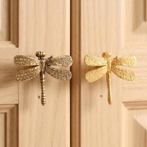 May include: Two dragonfly-shaped cabinet knobs made of metallic brass. Each knob features detailed, textured wings with a grid-like pattern and a segmented body. The knobs are mounted on a light-colored wooden cabinet door with vertical panels.