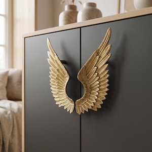 May include: A pair of gold-colored metal cabinet handles shaped like detailed angel wings. The handles are mounted on the two doors of a dark gray cabinet with a light wood-toned frame, featuring intricate feather textures on each wing.
