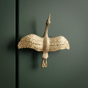 May include: A gold-colored metal cabinet knob shaped like a bird in flight with wings spread wide. The bird features detailed feather textures across its wings and body, with its head pointed upward and feet positioned at the bottom of the design.