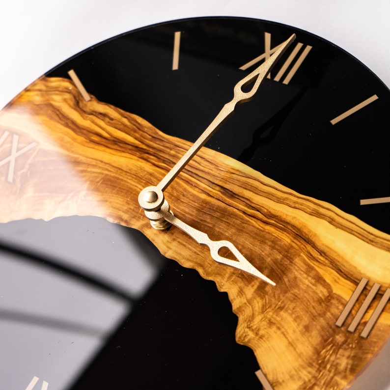 May include: A black and wood clock face with gold Roman numeral hour markers and gold hands. The clock face is made of epoxy resin and olive wood.