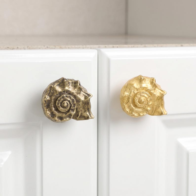 Two shell-shaped copper cabinet handles mounted on glossy white cabinet doors, shown in two styles/finishes: one darker antique-style shell knob and one polished gold shell knob. Decorative coastal cabinet hardware with a seashell design, ideal for beach house decor, nautical interiors, dressers, drawers, wardrobes, bathroom vanities, and kitchen cabinets.