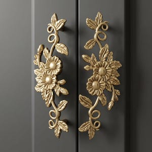 May include: Two gold-toned metal cabinet handles featuring an intricate floral design with embossed leaves, swirling vines, and blooming flowers. The handles are mounted vertically on a dark gray cabinet door, showcasing a metallic finish.