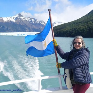May include: A woman in a blue jacket and sunglasses holds an Argentinian flag while standing on a boat. The flag is waving in the wind and the mountains and glacier are visible in the background.
