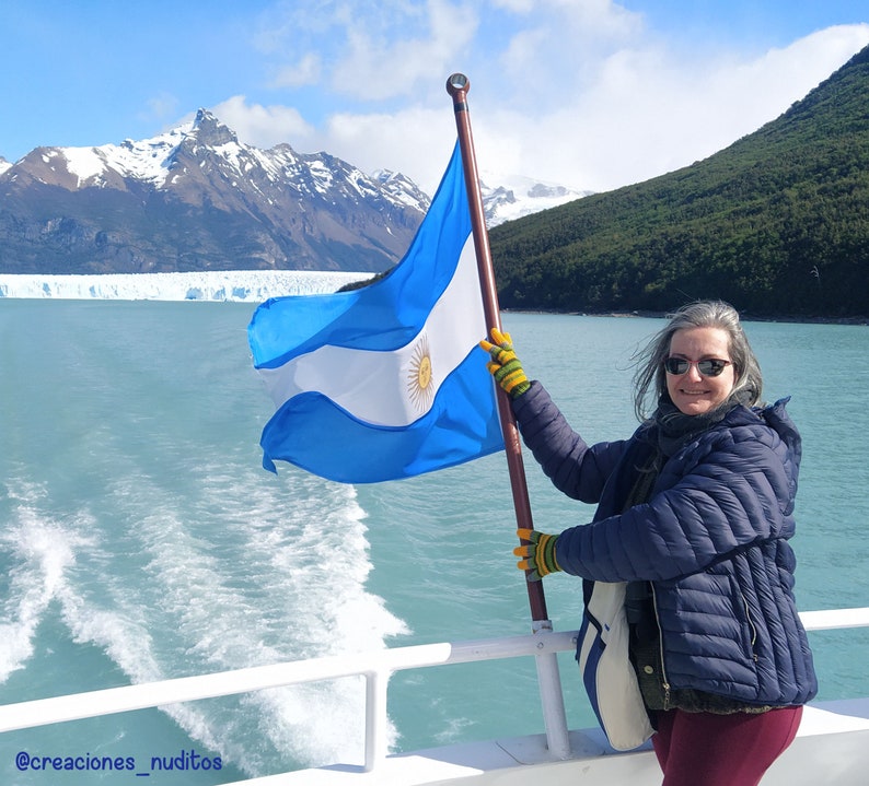 May include: A woman in a blue jacket and sunglasses holds an Argentinian flag while standing on a boat. The flag is waving in the wind and the mountains and glacier are visible in the background.