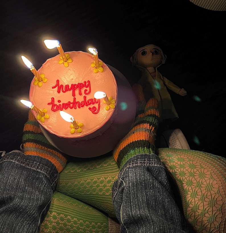 May include: A pink birthday cake with lit candles and the words "Happy Birthday" written in pink frosting. The cake is sitting on a table with a person wearing green and black striped socks and blue jeans.