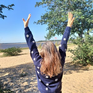 May include: A person with long brown hair wearing a blue sweater with white stars, standing with arms raised in the air on a sandy beach with a body of water in the background.
