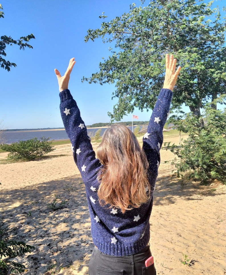 May include: A person with long brown hair wearing a blue sweater with white stars, standing with arms raised in the air on a sandy beach with a body of water in the background.