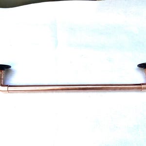 May include: A copper-colored metal towel bar with black circular mounting plates on each end. The bar is made of copper pipes and fittings, showcasing an industrial design. The bar is approximately 24 inches long.