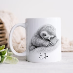 May include: A white ceramic mug featuring a detailed black and white illustration of a sleeping sloth resting on a branch. Below the sloth, the name Ella is printed in a black cursive script font. The mug has a standard handle on the left side.