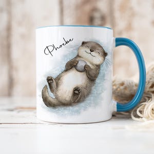 Pu&ograve; includere: Una tazza in ceramica bianca con manico e bordo blu, caratterizzata da un'illustrazione ad acquerello di una lontra marrone che galleggia sulla schiena tenendo una piccola pietra grigia. Il nome Phoebe &egrave; stampato in corsivo nero sopra.