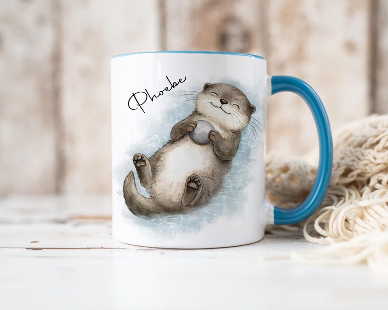 Pu&ograve; includere: Una tazza in ceramica bianca con manico e bordo blu, caratterizzata da un'illustrazione ad acquerello di una lontra marrone che galleggia sulla schiena tenendo una piccola pietra grigia. Il nome Phoebe &egrave; stampato in corsivo nero sopra.