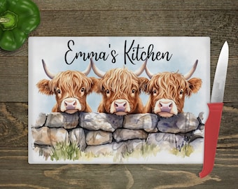 Può includere: Un tagliere rettangolare in vetro con un'illustrazione ad acquerello di tre mucche Highland marroni che spuntano da un muro di pietra grigia. Il testo Emma's Kitchen è stampato in nero sopra le mucche. È incluso un coltello con manico rosso.