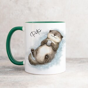 Pu&ograve; includere: Una tazza in ceramica bianca con manico e interno verde scuro. La tazza presenta un'illustrazione in stile acquerello di una lontra marrone che galleggia sulla schiena tenendo una piccola pietra grigia. Il nome Peter &egrave; scritto in nero sopra.