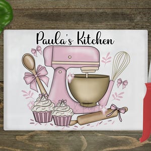 May include: A rectangular chopping board featuring a pink stand mixer, wooden spoon, whisk, rolling pin, and cupcakes. The words "Paula's Kitchen" are written in black script. A red-handled knife is on the right.