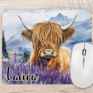 May include: A rectangular mouse mat with a watercolour painting of a Highland cow with long, shaggy, light brown fur, set against a backdrop of lavender and mountains. The name "Laura" is written in cursive.