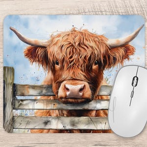 May include: A mouse mat with a watercolour illustration of a Highland cow. The cow has long, reddish-brown fur and large horns, looking over a wooden fence. A white computer mouse is positioned to the right of the mouse mat.