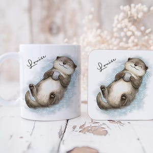 Pu&ograve; includere: Una tazza in ceramica bianca e un sottobicchiere quadrato, entrambi con un'illustrazione ad acquerello di una lontra marrone che galleggia sulla schiena tenendo una piccola pietra grigia. Il nome Louise &egrave; scritto in corsivo nero sopra l'animale.