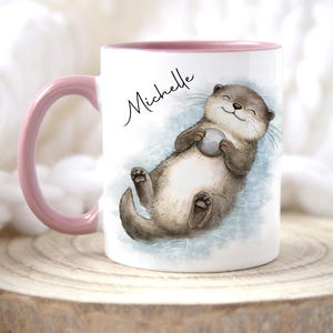 Pu&ograve; includere: Una tazza in ceramica bianca con manico e bordo interno rosa. La tazza presenta un'illustrazione stampata di una lontra marrone che galleggia sulla schiena tenendo una piccola pietra grigia. Il nome Michelle &egrave; stampato in nero sopra la lontra.
