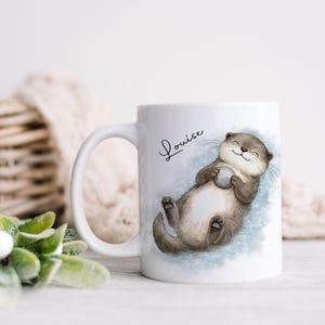 Pu&ograve; includere: Una tazza in ceramica bianca con l'illustrazione di una lontra marrone che galleggia sulla schiena tenendo una piccola pietra grigia. Il nome Louise &egrave; stampato in corsivo nero sopra la lontra, su uno sfondo ad acquerello blu tenue.